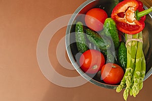 Vegetables lie in a metal bowl: tomatoes, asparagus, cucumbers, red bell peppers. on a wooden board and brown background