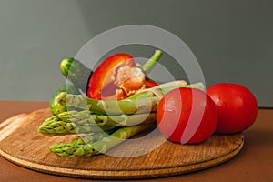 Vegetables lie on a wooden board: tomatoes, asparagus, cucumbers, red bell peppers. brown, dark gray background. place