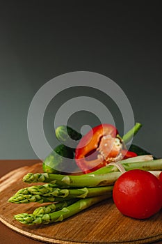 Vegetables lie on a wooden board: tomatoes, asparagus, cucumbers, red bell peppers. brown, dark gray background. place