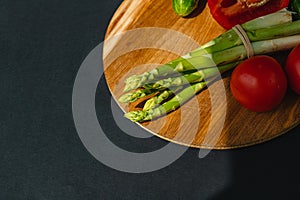 Vegetables lie on a wooden board: tomatoes, asparagus, cucumbers, red bell peppers. brown, dark gray background. place