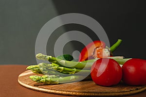 Vegetables lie on a wooden board: tomatoes, asparagus, cucumbers, red bell peppers. brown, dark gray background. place