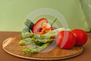 Vegetables lie on a wooden board: tomatoes, asparagus, cucumbers, red bell peppers. brown, light green background. place