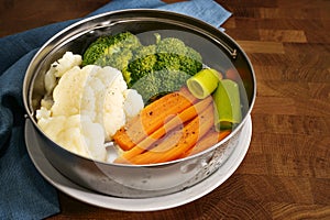 Vegetables such as carrots, broccoli and cauliflower in a metal food steamer basket on a dark rustic wooden table, cooking method