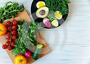 Vegetarian food on a light background, fresh vegetables on a cutting board and greens on a plate with avocado and egg, top view