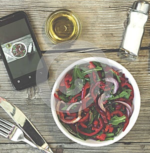 Vegetarian lunch. Salad of fresh vegetables, greens and red sweet peppers with olive oil. On the smartphone screen, a lunch displa