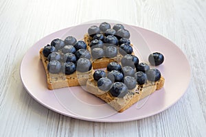 Vegetarian toasts with peanut butter, blueberries and chia seeds on a pink plate over white wooden surface, top view. Healthy eati