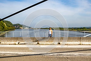 Vehicular road in poor condition on Lanaye cable-stayed bridge, woman hiker standingert canal, two boats sailing, trees,