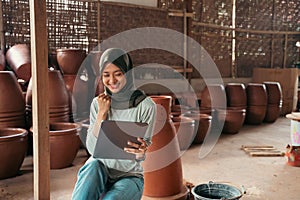 veiled asian woman smiling using tablet while sitting between pottery