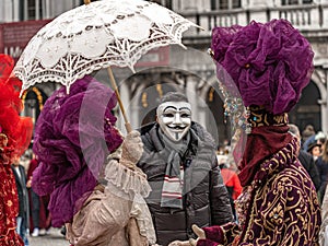 Venice Carnival. A man in Guy Fawkes mask is posing with a costumed masked woman with a parasol and a costumed masked man