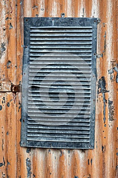 A vent in the wall of a corrugated metal building with peeling orange paint