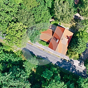 Vertical aerial view of a house with a saddle roof and red roof tiles in a wooded area next to an asphalted grey country road