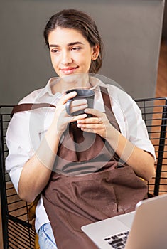 Vertical angle of beautiful caucasian barista woman looks blankly outside coffee shop while relaxing in coffee break in front of