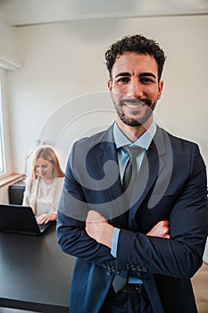 Vertical arms crossed businessman smiling and looking at camera standing at corporate office. Clerk male staring front