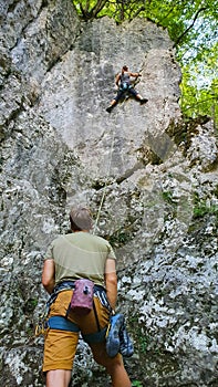 VERTICAL: Athletic female rock climber is being belayed by unrecognizable man.