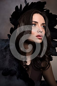 Vertical beauty portrait in dark tones. Beautiful young woman in a cloud of smoke with black feathers in her hair
