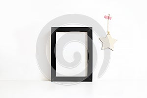 Vertical black blank wooden frame mockup on white table. Wooden star decoration hanging on the wall. Styled stock