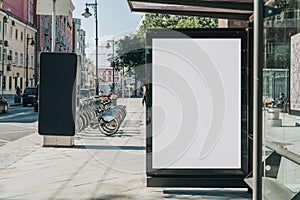 Vertical blank billboard at bus stop on city street. In background buildings, road. Mock up. Poster next to roadway.