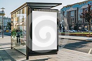 Vertical blank billboard at bus stop on city street. In background buildings, road. Mock up. Poster next to roadway.