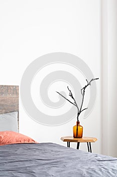 Vertical close-up of glass orange vase with a tree branch standing on a simple wood table next to the bed