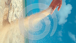 VERTICAL CLOSE UP: Grains of white sand fly away in the wind from unrecognizable playful woman`s hand while enjoying a relaxing af