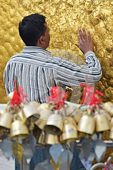 Vertical close up view of Pilgrim carefully pasting gold foils onto golden rock at the Kyaiktiyo Pagoda with row of small bells