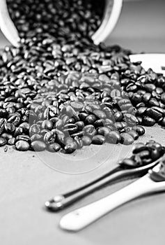 Vertical closeup grayscale of coffee beans pouring from a wooden mug on a table with spoons