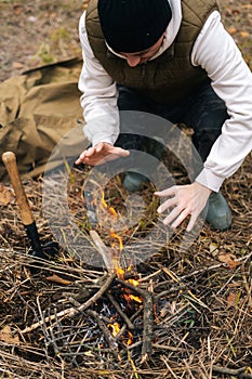 Vertical cropped shot of frozen traveler man warming hands over fire at outdoors on overcast cold day.