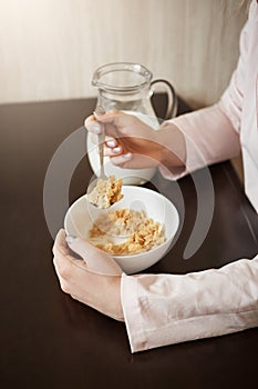 Vertical cropped shot of woman sitting in kitchen holding spoon while eating bowl of cereals with milk, having healthy