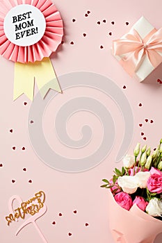 Vertical flat lay composition with rosette best mom ever, gift box, bouquet of flowers on pink background with confetti. Happy