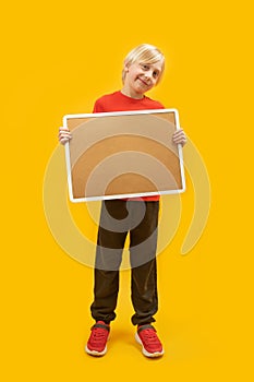 Vertical frame full-length portrait of blond boy with cortical board in his hands, yellow background. Place to advertise. Copy