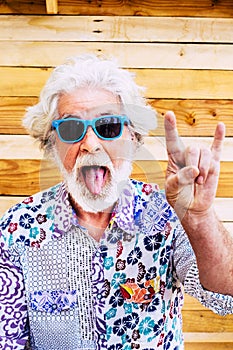 Vertical front portrait of old mature man with tongue and devil gesture. Fun senior people with colorful clothes and crazy