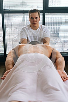 Vertical front view of professional male masseur with strong hands massaging shoulder of muscular sports man lying on