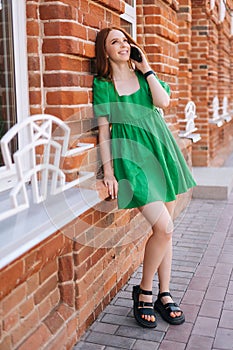 Vertical full length portrait of laughing young woman talking on mobile phone standing leaning on old red stone wall at