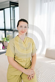Vertical full length portrait of middle-aged friendly female masseuse therapist in uniform sitting on white couch on