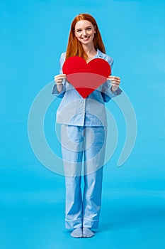 Vertical full-length portrait redhead elegant woman in cute pyjama, holding big red heart sign, valentines day gift and