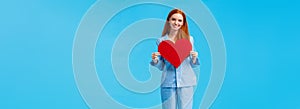 Vertical full-length portrait redhead elegant woman in cute pyjama, holding big red heart sign, valentines day gift and