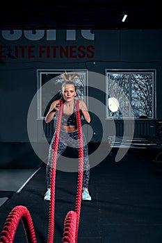Vertical full length shot of an athletic fitness woman doing functional training exercise with battle ropes at the gym