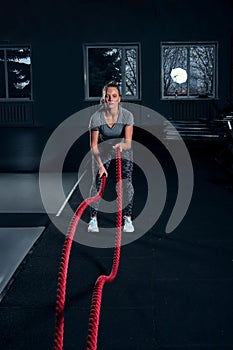 Vertical full length shot of an athletic fitness woman doing functional training exercise with battle ropes at the gym