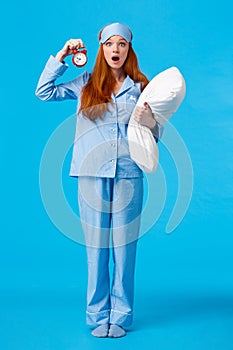 Vertical full-length shot shocked and startled cute redhead female in sleeping mask and pyjama, holding pillow and alarm