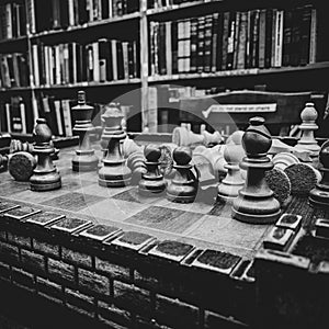 Vertical grayscale closeup of the chess pieces on the board against the bookcase. Selected focus.