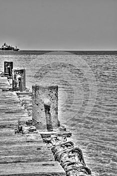 Vertical grayscale of an old wooden dock of a lake, rusty poles on sides and a yacht on the horizon