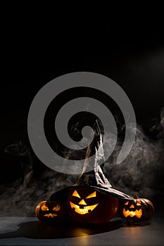 Vertical Halloween card. Witch hat on a pumpkin with carved creepy grimaces on a black background in the fog. Jack-o