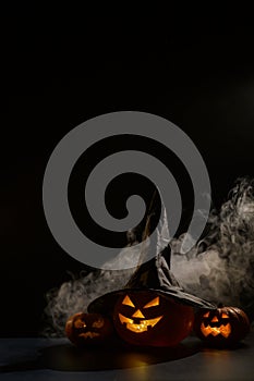 Vertical Halloween card. Witch hat on a pumpkin with carved creepy grimaces on a black background in the fog. Jack-o