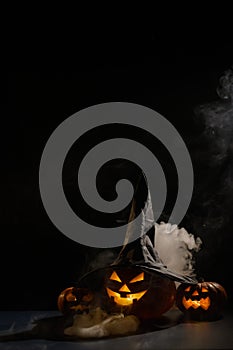 Vertical Halloween card. Witch hat on a pumpkin with carved creepy grimaces on a black background in the fog. Jack-o