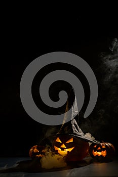 Vertical Halloween card. Witch hat on a pumpkin with carved creepy grimaces on a black background in the fog. Jack-o