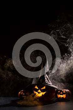 Vertical Halloween card. Witch hat on a pumpkin with carved creepy grimaces on a black background in the fog. Jack-o