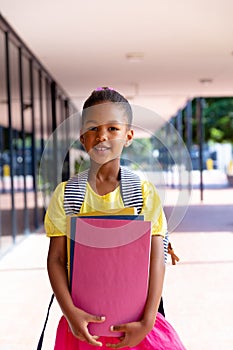 Vertical of happy biracial schoolgirl with school bag holding books outside school, with copy space