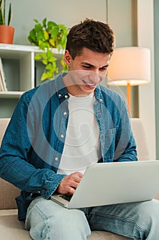 Vertical. Happy freelancer man working at home with a laptop on a sofa. Young guy smiling and browsing on internet using