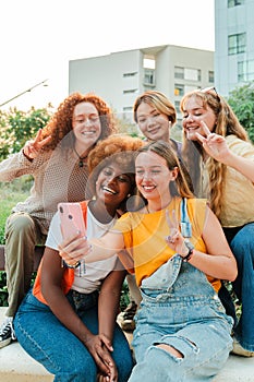 Vertical. Happy multiracial young women looking at camera with a smart phone enjoying together sitting outside. Funny