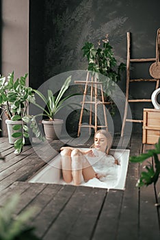 Vertical image of beautiful blonde young woman taking bath with bubbles in bathroom interior with plenty green plans